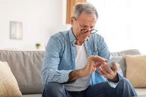 An older man wearing glasses is sitting on a couch while holding a smartphone and seems to be texting or browsing something on it.