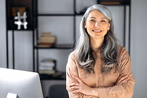 A woman with gray hair is standing with her arms crossed in an office. Behind her, there is a white desk with a monitor, a shelf with books, and a white wall with a blurry white and black hashtag symbol.