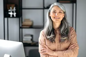 A woman with gray hair is standing with her arms crossed in an office. Behind her, there is a white desk with a monitor, a shelf with books, and a white wall with a blurry white and black hashtag symbol.