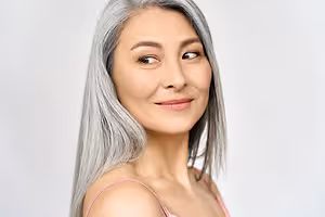 Portrait of a mature Asian woman with gray hair and a pink top, smiling in a white room