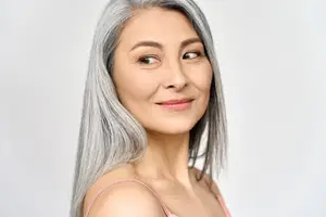Portrait of a mature Asian woman with gray hair and a pink top, smiling in a white room