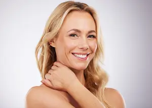 A woman with blonde hair smiles and poses with one hand on her neck
