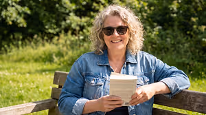 A woman in a denim jacket and sunglasses sits on a bench and reads a book