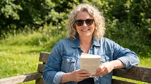 A woman in a denim jacket and sunglasses sits on a bench and reads a book
