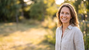 A smiling woman stands in a park on a sunny day, surrounded by greenery.