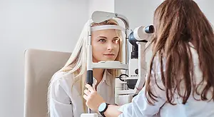 A woman wearing a white shirt is sitting in a chair and being examined by a doctor wearing a white shirt and a watch with an ophthalmoscope.
