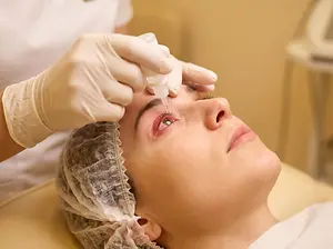A woman is lying on a chair while a beautician is applying a serum on her forehead with a dropper.