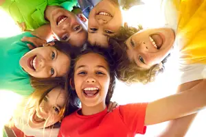 A group of children in a circle smiling at the camera, with one child in the middle wearing a red shirt, and another child on the right wearing a yellow shirt