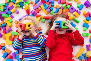 Two young children lying on a floor filled with colorful building blocks, they are holding a blue block in their hands and smiling at the camera.