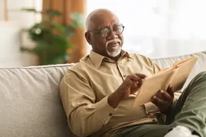 A smiling elderly man wearing glasses and a beige shirt is sitting on a couch, reading a book and looking at the camera.