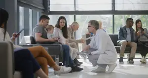 A group of people are sitting in a room with glass walls, and one man is holding a paper while bending down. Some people are sitting on the chairs, and some are standing. Some of them are looking at their cell phones, while others are looking at the man. There are trees outside the glass walls. The room has a bright light from the outside. 
