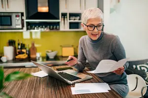 A woman in glasses sitting at a kitchen table, working on a laptop and reading a document.