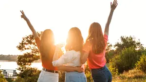 Three young women standing together on a hillside overlooking a lake during sunset with their arms raised in the air