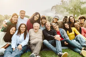 A group of people are sitting on the grass and seem to be posing for a photo in a park. They are all smiling and seem to be happy. Some of them are wearing glasses. They are sitting on the grass with some trees behind them. The sky is white and clear.