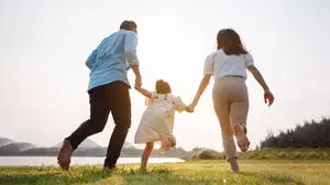 A family of three, consisting of a man, a woman, and a little girl, are walking together on a grassy field with a lake and mountains in the background during a sunny day.