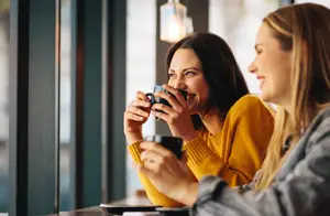 Two young women sitting at a bar drinking coffee and laughing