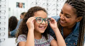 A young girl is smiling as she tries on a pair of glasses in a store with a woman looking at her. Behind her, another woman is visible in the mirror.