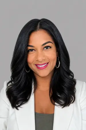 A smiling woman with black hair wearing a white blazer, standing against a gray background, posing for a photo