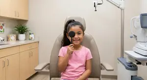 A young girl is sitting in an optometrist's chair, holding an eye patch over her left eye, and smiling while looking forward. Behind her, a sink with a potted plant on the counter, cabinets, and various medical equipment are visible in the background.