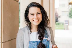A woman smiling and posing for a photo with curly hair and a gray cardigan and denim overalls