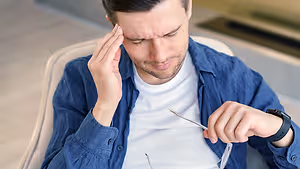 A man is sitting in a chair, holding his head with his left hand, and seems to be adjusting his eyeglasses with his right hand. He is wearing a white t-shirt and a blue jacket. The man is looking down, possibly concentrating on adjusting his eyeglasses. Behind him is a blurry background, which seems to be a room with a glass wall and a white floor.
