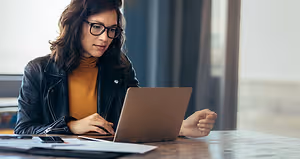 A woman is sitting at a desk, working on a laptop and holding a pen in her right hand. She is wearing glasses and a black jacket. There is a book, a cell phone, a pen, and a notebook on the desk. Behind her is a window with a blue curtain. The light from outside is illuminating the room.