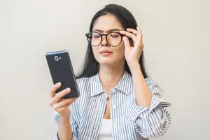 A woman with glasses squinting and holding a phone with one hand on her forehead