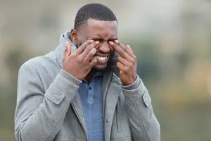 A man standing outside in a park, wearing a grey hoodie, with his hands covering his eyes and smiling