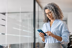 A woman in a striped shirt and glasses is looking at her phone in an office building with glass walls.