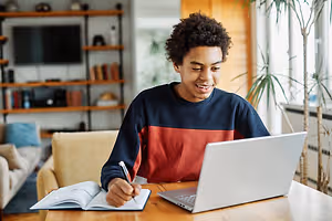 A young man is smiling and writing on a notepad while using a laptop on a wooden table in a room.