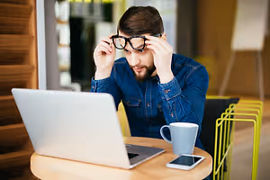 A man in a denim shirt adjusts his glasses while looking at a laptop on a table with a cup and a phone.