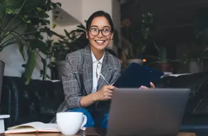 A woman in a suit holding a pen and a folder while sitting on a couch in a room with plants and a laptop.