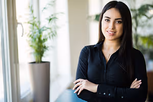 A woman with long black hair and a black shirt is standing with her arms crossed in an office room with a glass window and a potted plant