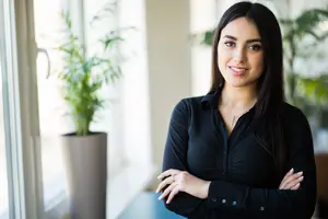A woman with long black hair and a black shirt is standing with her arms crossed in an office room with a glass window and a potted plant