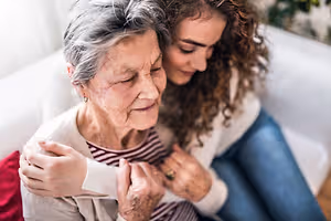 An older woman and a young woman are sitting on a couch, and the young woman is hugging the older woman. The older woman is wearing a white and black striped shirt, and the young woman is wearing a white long-sleeve shirt and blue jeans. Behind them, there is a blurry image of a white couch, and there is a plant in the corner of the room.