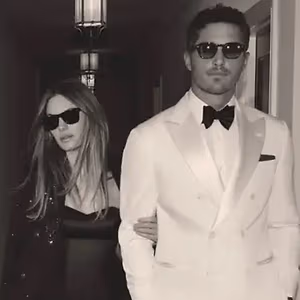 A man and woman dressed in formal attire, standing in a corridor, with the man wearing a bow tie and the woman wearing sunglasses