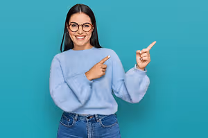 A woman wearing a blue sweater and glasses is pointing her finger at a blue background while smiling.