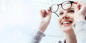 A smiling woman wearing glasses is holding them up to her face in a room.