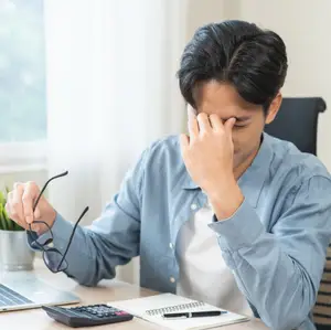 Stressed man sitting at a desk looking at a calculator with his hand on his forehead.