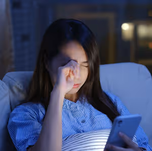 A young woman sitting on a couch and looking at her phone while holding her nose and closing her eyes.