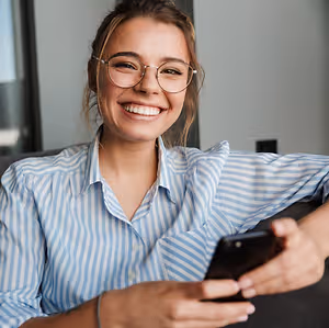A woman in glasses and a blue and white striped shirt is smiling while looking at her phone.