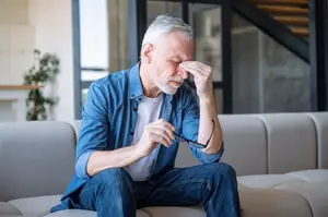 An older man with white hair and a white beard sitting on a couch looking stressed with his hand on his forehead.