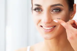 A woman with green eyes and white nails is smiling and placing a contact lens on her eye