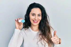 A woman is holding a pair of contact lenses in her hand and smiling