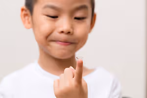 A boy wearing a white shirt is holding a contact lens and smiling at the camera.