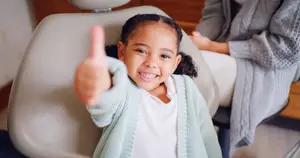 A young girl gives a thumbs up while sitting in a dentist chair with a woman behind her, both smiling.