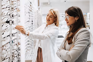 Two women standing next to a wall of glasses in a store, the woman on the left is holding a pair of glasses and smiling while the woman on the right is looking at her with a smile on her face.