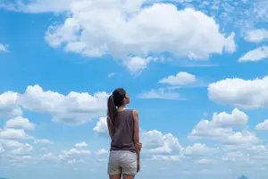 A woman wearing a brown tank top and white shorts is standing on a hill looking at the sky with her hands on her hips