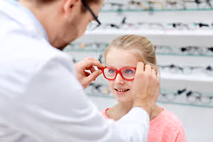 A doctor is adjusting a pair of red glasses on a girl's face.