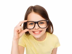 A young girl with brown hair is smiling and holding a pair of black glasses up to her face.